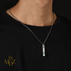 Puede incluir: Un collar plateado con un colgante rectangular. El colgante es blanco con dos rayas diagonales azules. El collar se lleva sobre una camiseta negra.