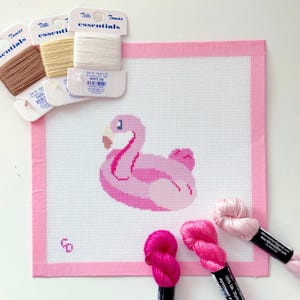 May include: A square needlepoint canvas featuring a pink flamingo design with a pink border. Included are three spools of Tilli Tomas Essentials silk thread in white, cream, and brown, plus three skeins of pink silk embroidery floss in varying shades.