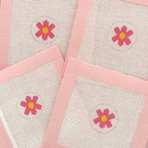 May include: Four square coasters with a pink border and a white mesh center. Each coaster features a cross-stitched pink flower with a yellow center. The coasters are arranged in a slightly overlapping pattern.