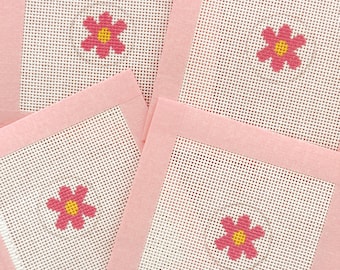 May include: Four square coasters with a pink border and a white mesh center. Each coaster features a cross-stitched pink flower with a yellow center. The coasters are arranged in a slightly overlapping pattern.
