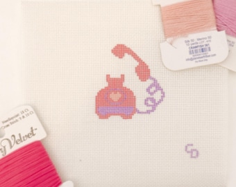 May include: A cross-stitch design of a pink and purple telephone on a white canvas. Spools of pink and purple thread are in the background. The text "Title Tomas essentials" is visible on the thread spools.