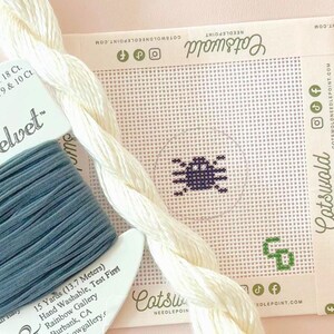 Puede incluir: Un lienzo cuadrado para bordar con un diseño de pequeña araña morada, acompañado de un carrete de hilo de terciopelo azul y una hebra de lana color crema. El lienzo incluye el logotipo y la dirección web de Cotswold Needlepoint.