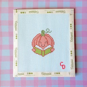 May include: A square needlepoint canvas featuring a light blue grid background and a central cross-stitch design of a pink pumpkin reading a green book. The edges are bound with beige tape printed with the text Catswold and social media icons.