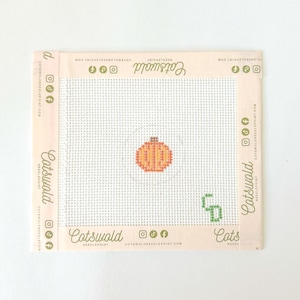 Könnte beinhalten: Ein quadratisches Stickleinen mit weißem Gittergewebe, das in der Mitte ein kleines, gesticktes orangefarbenes Kürbismotiv und in der unteren rechten Ecke ein grünes Monogramm aufweist. Die Ränder sind mit hellrosa Klebeband mit der Aufschrift Cotswold eingefasst.