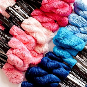 Pode incluir: Uma coleção de fios de seda coloridos em tons de rosa, azul e marinho. Cada meada é rotulada com o nome da marca e a composição da fibra. A imagem mostra uma variedade de cores, perfeitas para bordados ou outros trabalhos têxteis.