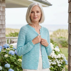 Op de afbeelding: Een lichtblauwe cardigan met een ronde hals en parelknopen. De geribbelde gebreide trui wordt over een witte top gedragen. De achtergrond omvat blauwe en witte bloemen, een houten huis en uitzicht op de zee.