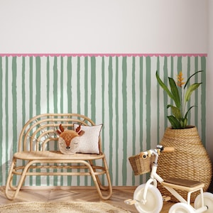 Puede incluir: Una habitación infantil con una pared a rayas verdes y blancas y un borde rosa festoneado. Un banco de mimbre natural sostiene un cojín con forma de ciervo. Una cesta tejida contiene una planta y un pequeño triciclo blanco con una cesta de mimbre.