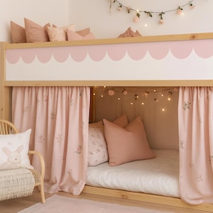 Op de afbeelding: Een licht houten stapelbed met roze geschulpte rand en gordijnen. Het bed is versierd met roze kussens en een bloemenslinger. Een rotan stoel met een konijnenkussen staat voor het bed.