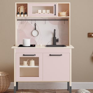 May include: A pink and white wooden play kitchen with a microwave, sink, and stove. The kitchen has a light pink cabinet and a white countertop. Accessories include a pan, a teapot, and a white cup. The kitchen is designed for children.