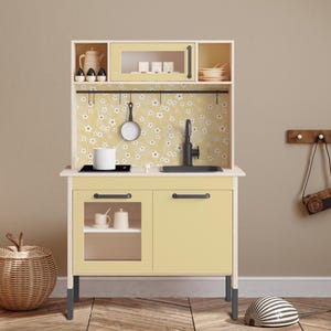 Op de afbeelding: Een gele houten speelkeuken met een zwarte gootsteen, fornuis en kastdeuren. De keuken heeft een witte betegelde achterwand met een bloemenpatroon en een klein plankje boven de gootsteen. De keuken staat op zwarte poten en heeft een klein raam in de kastdeur.