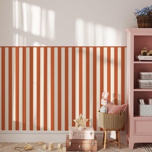 Op de afbeelding: Een kinderkamer met oranje en witte gestreepte behang. Een roze kast bevat speelgoed en decoratieve items. Een rieten stoel bevat een pop en een kussen. Koffers en een stervormige decoratie zijn op de vloer gestapeld.