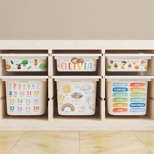 May include: Six white storage bins with colourful illustrations and text. The bins are labelled "Vegetables", "Fruits and Berries", "Numbers", "Weather", "Month of the Year", and "Little Baby Olivia". The bins are inside a light wood shelf.