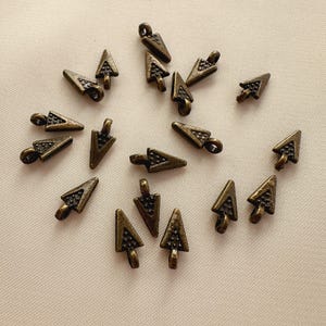 May include: A collection of small, bronze-colored arrow charms with a loop at the top for attaching to jewelry or other crafts. The arrows have a textured surface and a vintage aesthetic.