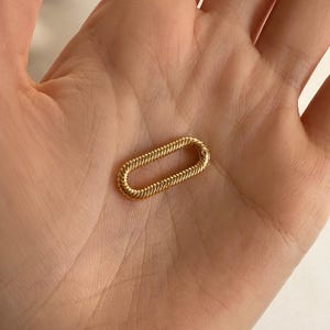 Peut inclure: Un maillon de bijou doré de forme ovale avec un motif tressé texturé. Le maillon est présenté sur un fond neutre, soulignant son éclat métallique et ses détails complexes. Ce bijou peut être utilisé dans les colliers, bracelets ou autres bijoux.