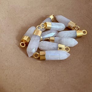 Könnte beinhalten: Eine Sammlung hellgrauer Kristallanhänger mit goldfarbenen Kappen und Schlaufen. Die Anhänger sind sechseckig und leicht unterschiedlich groß. Der Hintergrund ist einfarbig warmbraun.
