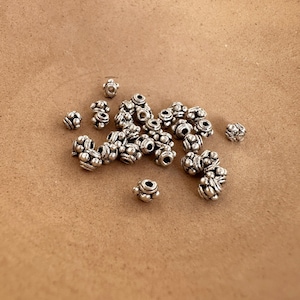 Peut inclure: Un tas de perles en métal argenté avec un motif décoratif. Les perles sont rondes et présentent un motif en relief de petits cercles.