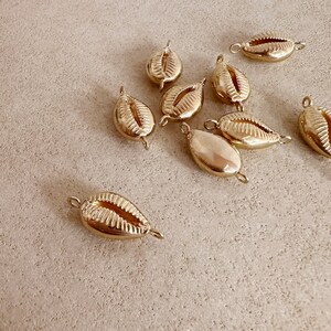 May include: A selection of gold-coloured cowrie shell charms. Each charm has a loop at each end for attaching to jewellery. The charms are scattered on a neutral-toned surface, highlighting their polished, reflective finish. These charms are suitable for creating unique necklaces, bracelets, and earrings.