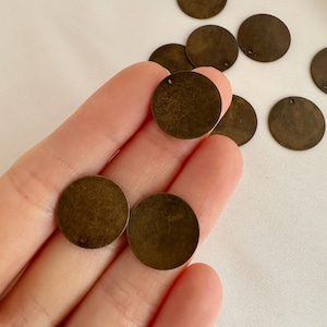以下が含まれることがあります： 小さな丸いブロンズ色の金属ディスクのコレクション。使い古したような仕上がりで、各ディスクの端近くに小さな穴があります。ディスクは白い表面に散らばっており、一部は手で持たれています。
