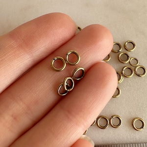 Op de afbeelding: Een close-up van een hand die een stapel kleine, goudkleurige springringen vasthoudt. De ringen zijn ongeveer 5 mm in diameter en zijn perfect voor het maken van sieraden.