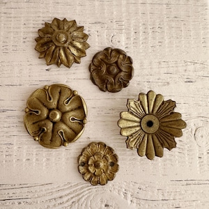 Op de afbeelding: Vijf antieke bloemvormige decoratieve elementen van messing. De elementen variëren in grootte en ontwerp, met gedetailleerde bloemblad- en middenpatronen. Het messing heeft een warme, verouderde patina en de stukken zijn gerangschikt op een verweerd wit houten oppervlak.