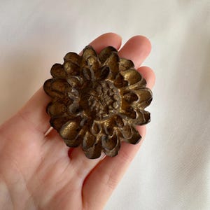 May include: A detailed, antique-style bronze-coloured metal flower. The flower has multiple layers of petals and a textured centre. The metal has a slightly aged appearance, with hints of gold and brown. The flower is likely a decorative element.