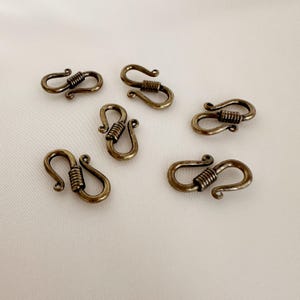 Puede incluir: Varios cierres en forma de S de color bronce antiguo. Cada cierre presenta ganchos curvos en ambos extremos, conectados por una sección de alambre enrollado. Los cierres están dispuestos sobre una superficie blanca, mostrando su diseño detallado y estética vintage.