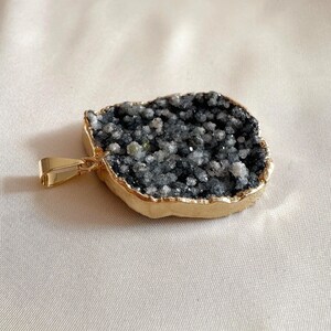Peut inclure: Pendentif avec une pierre druzy gris foncé ornée de cristaux scintillants, sertie dans une lunette dorée. Le pendentif est doté d'une bélière dorée pour l'attacher à une chaîne. La pierre a une texture naturelle rugueuse.