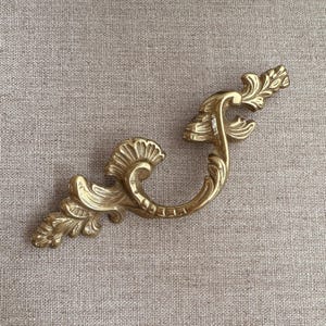 May include: Ornate gold-coloured furniture handle with a curved design. The handle features detailed leaf and scrollwork patterns, adding a touch of vintage elegance. The handle is approximately 15 cm long.