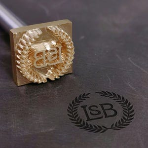 以下が含まれることがあります： 金色の金属スタンプ。イニシャル「LSB」が月桂樹のデザインの中にあります。スタンプは暗い表面に押し付けられ、一致する印象を残します。スタンプは正方形のヘッドと長いハンドルを持っています。