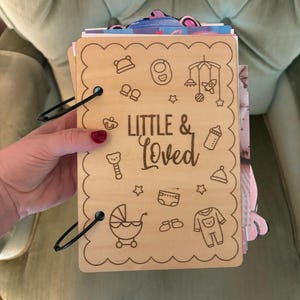 May include: A rectangular light-colored wooden plaque with scalloped edges, bound by two black metal rings. The front features the engraved text "LITTLE & LOVED" surrounded by icons of a baby hat, bib, mobile, pacifier, bottle, rattle, diaper, stroller, and onesie.