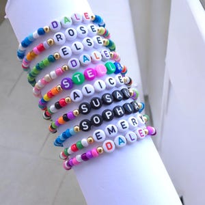 Könnte beinhalten: Ein Stapel aus zehn bunten Perlenarmbändern mit weißen und schwarzen Buchstabenperlen, die die Namen DALE, ROSE, ELSE, DALE, STELLA, ALICE, SUSAN, SOPHIA, EMERY und DALE bilden. Die Armbänder enthalten kleine goldfarbene Zwischenperlen.