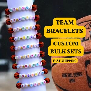 Puede incluir: Pulseras de equipo personalizadas con nombres y números, con cuentas temáticas de baloncesto. Las pulseras son blancas con cuentas de letras coloridas y pequeños acentos de baloncesto. La imagen también incluye una pelota de baloncesto y el texto "Team Bracelets, Custom Bulk Sets."