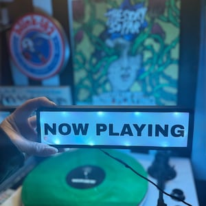 May include: A black rectangular LED sign with the words "NOW PLAYING" in white. The sign is held above a green vinyl record on a turntable. The background includes a record album and a circular logo.