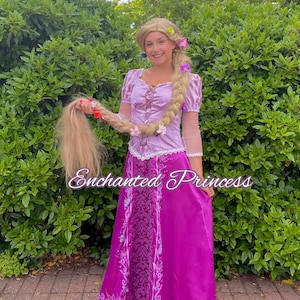 Könnte beinhalten: Eine Frau in einem lila Prinzessinnenkostüm mit einem langen blonden Zopf, der mit Blumen geschmückt ist. Das Kostüm umfasst ein Oberteil, einen langen Rock und eine weiße Spitzenborte. Der Text "Enchanted Princess" ist sichtbar.