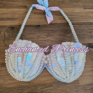May include: A white and iridescent mermaid-style bra top with pearl accents and a light blue ribbon tie. The bra top is decorated with a shell-like design and has a pearl trim.