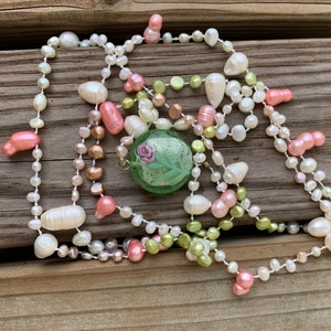May include: A multi-strand pearl necklace with a green glass pendant featuring a pink rose design. The necklace is made of various sizes and shapes of pearls in white, pink, and green.