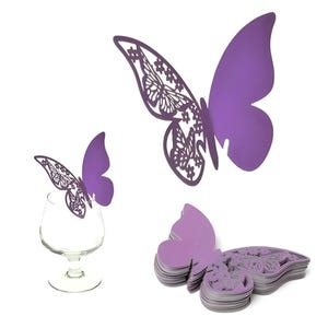May include: Purple butterfly-shaped decorations. One butterfly is perched on a wine glass, while others are displayed separately. Some butterflies have intricate cut-out wing designs. The butterflies are made of a solid material and are a vibrant purple color.