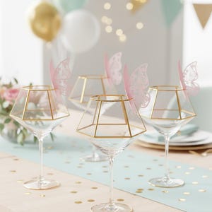 May include: Four clear martini glasses with gold geometric frames and pink butterfly accents. The glasses sit on a light blue table runner with gold confetti. Balloons and party decorations are in the background.