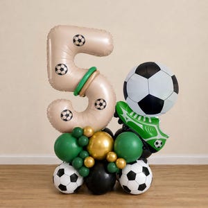 May include: A balloon arrangement featuring a large beige number five balloon decorated with soccer ball patterns, a round soccer ball balloon, and a green soccer cleat balloon. The base consists of green, gold, black, and soccer ball balloons.