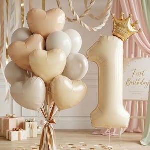 May include: A collection of cream and peach-coloured balloons, including heart-shaped and round balloons. A large number one balloon and a gold crown are also present. The scene is set for a first birthday celebration.