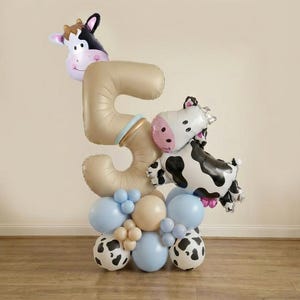 May include: A celebratory balloon display for a fifth birthday. The focal point is a large number 5 balloon in a neutral colour, decorated with cow-shaped balloons and a base of blue, beige, and cow-print balloons. A fun party decoration.