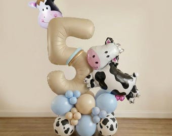 May include: A whimsical balloon arrangement for a fifth birthday. It features a large number 5 in a beige color, adorned with cow-shaped balloons and a base of blue, beige, and cow-print balloons. The arrangement is set against a neutral background.