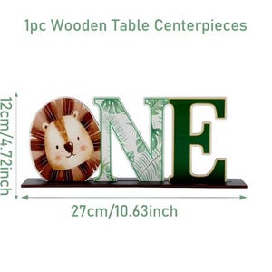 May include: A wooden table centerpiece spelling the word ONE, featuring a lion face for the letter O, a tropical leaf pattern on the letter N, and a solid dark green letter E. It measures 4.72 inches high and 10.63 inches wide.