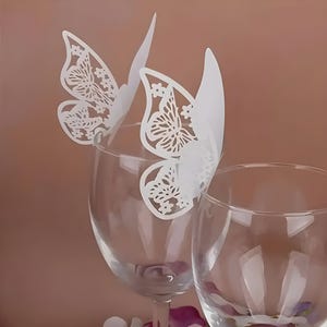 May include: Two clear wine glasses with white butterfly-shaped decorations. The butterfly wings are intricately cut with floral and geometric patterns. The background is a soft, muted brown.