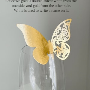 May include: A gold butterfly decoration with intricate cut-out details, perched atop a clear wine glass. The butterfly is double-sided, gold on one side and white on the other. Text on the image states that the white side is for writing a name.