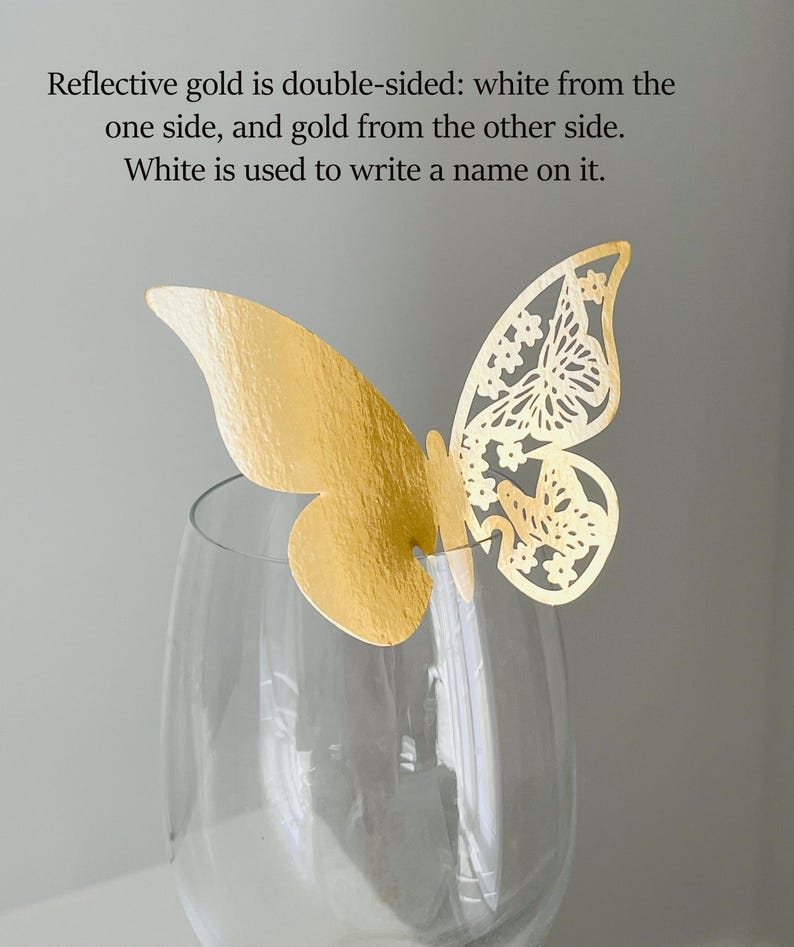 May include: A gold butterfly decoration with intricate cut-out details, perched atop a clear wine glass. The butterfly is double-sided, gold on one side and white on the other. Text on the image states that the white side is for writing a name.