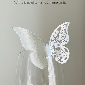 May include: A white, double-sided butterfly decoration with intricate cut-out details, perched on a clear wine glass. The text states the butterfly is reflective silver on one side and white on the other, suitable for writing a name.