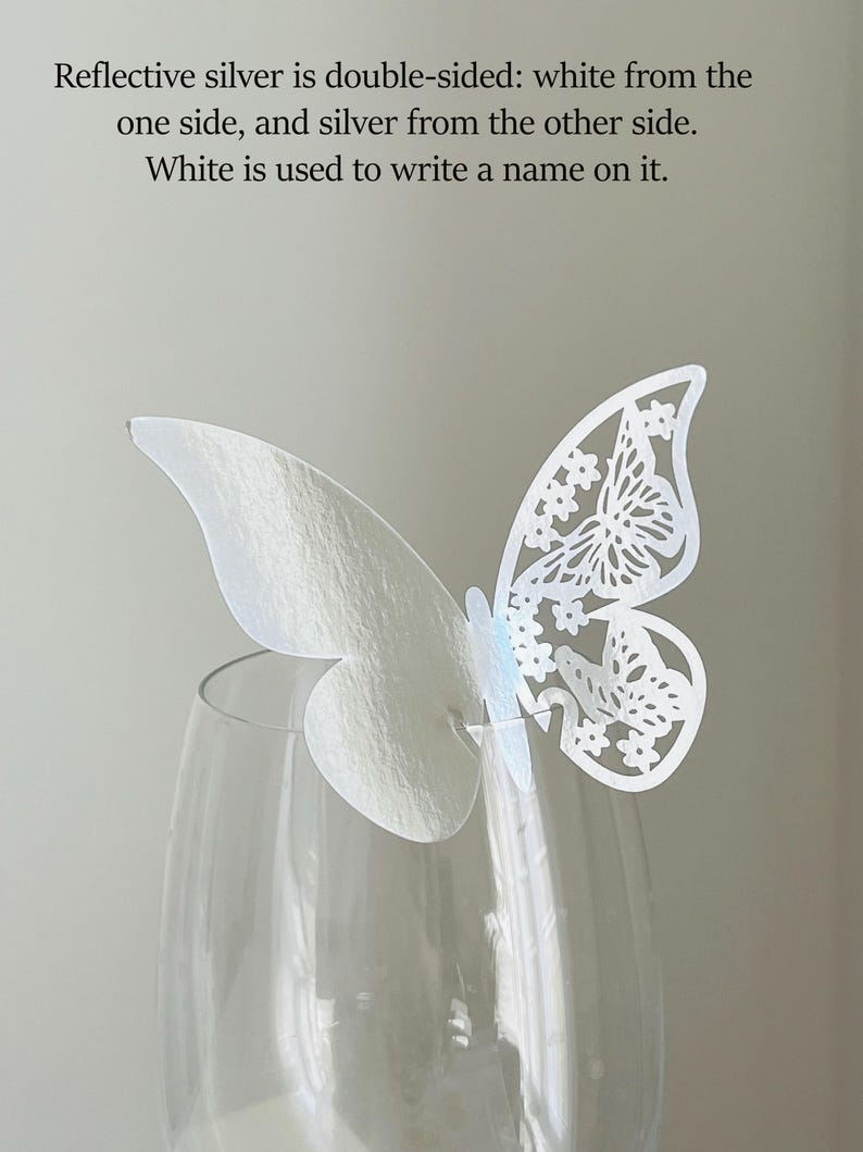 May include: A white, double-sided butterfly decoration with intricate cut-out details, perched on a clear wine glass. The text states the butterfly is reflective silver on one side and white on the other, suitable for writing a name.