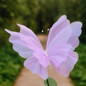 May include: A decorative butterfly ornament featuring translucent, light purple layered wings with delicate white vein detailing. The center body is adorned with a vertical row of small white beads, and two thin antennae extend upward with white tips.