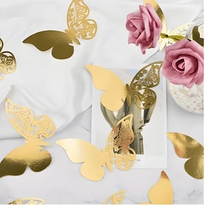 May include: Gold-colored butterfly decorations with intricate cut-out designs are scattered across a white surface. Pink roses and a decorative jar are also present, creating a delicate and elegant aesthetic. These butterflies are ideal for crafts and decor.